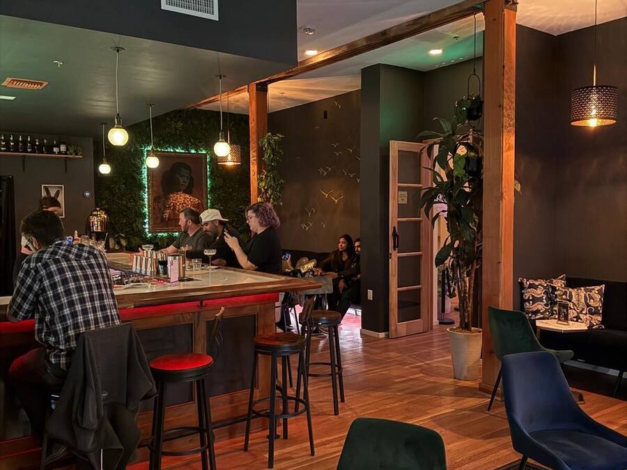 A modern bar area with wooden flooring, bar stools, and hanging pendant lights. There are seating areas with green upholstered chairs and a glass table with a menu.