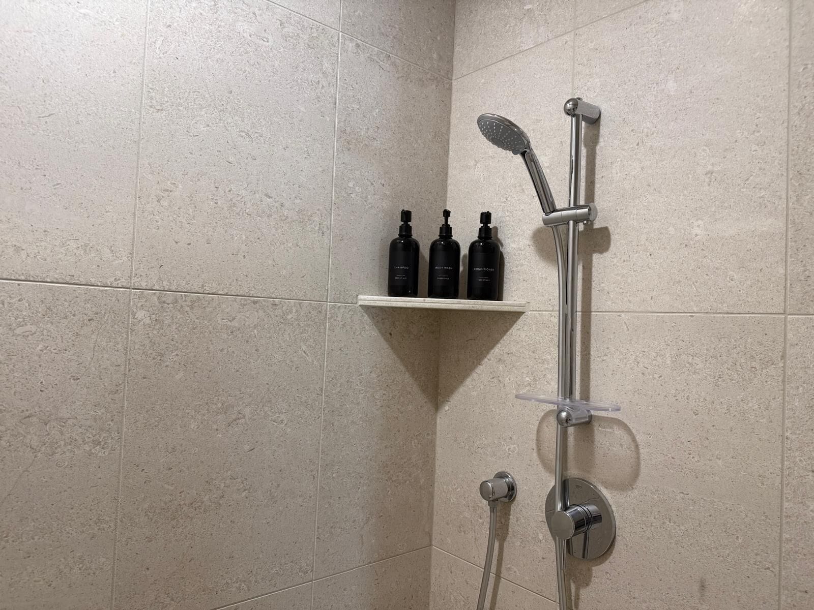 A shower area with a handheld showerhead and a wall-mounted faucet. There is a shelf with three bottles of shampoo, conditioner, and body wash.