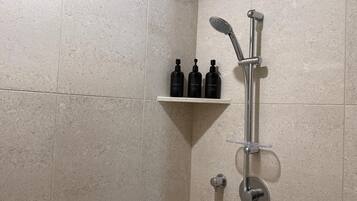 A shower area with a handheld showerhead and a wall-mounted faucet. There is a shelf with three bottles of shampoo, conditioner, and body wash.
