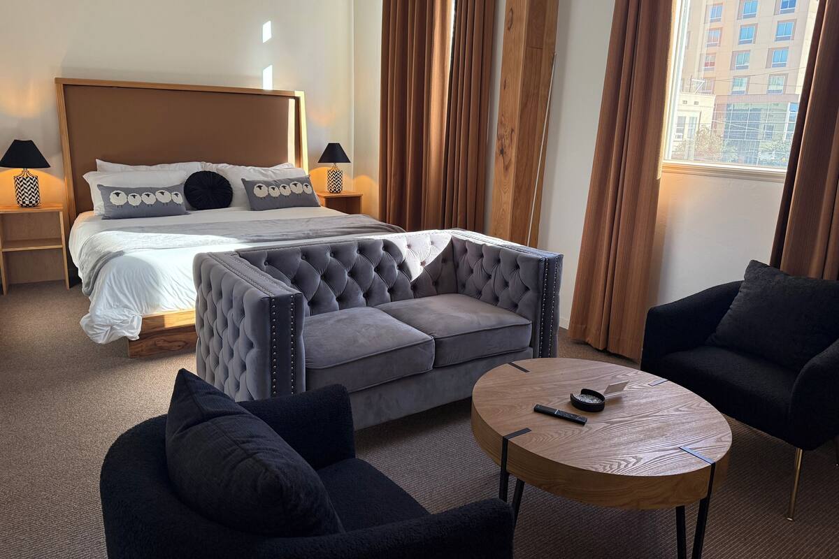 Hotellihuoneessa on sänky, harmaa, napitettulla kankaalla verhoiltu sohva, pyöreä puinen sohvapöytä ja kaksi nojatuolia.