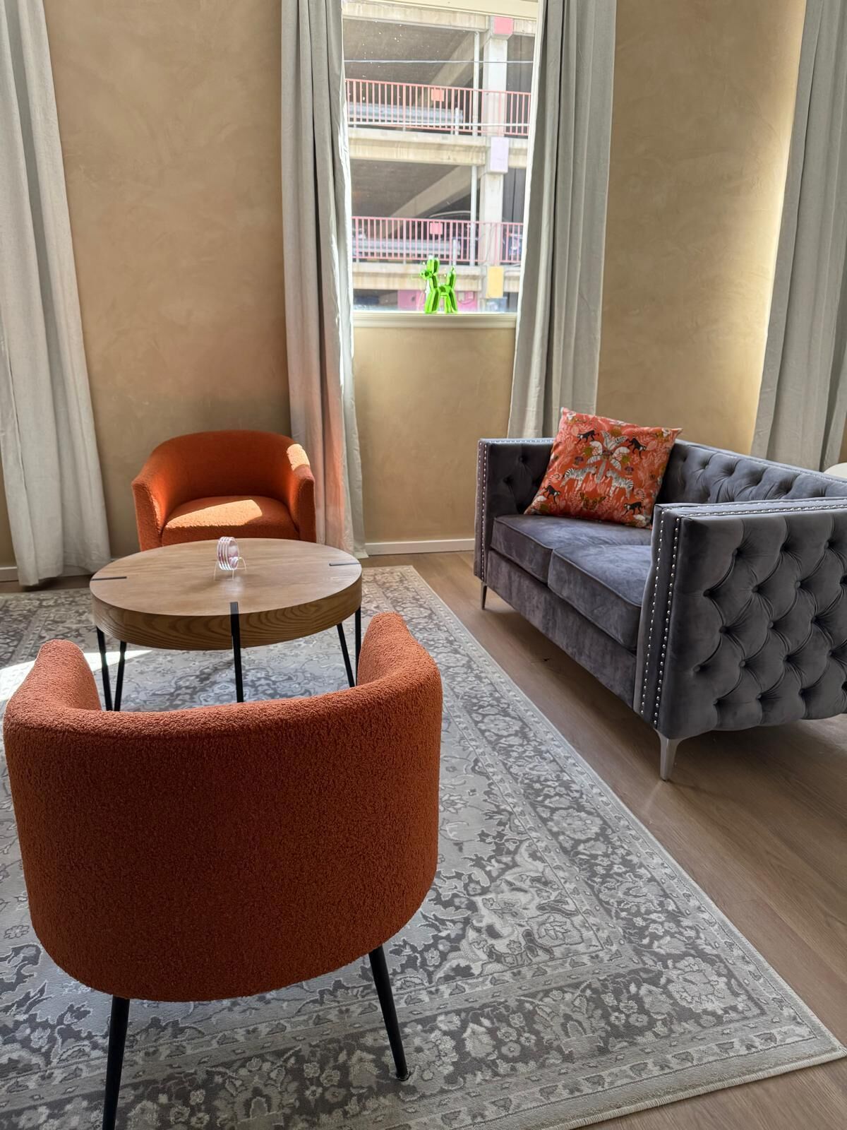 A living room with a round wooden table, two orange chairs, a grey tufted sofa with a floral pillow, and a patterned rug.