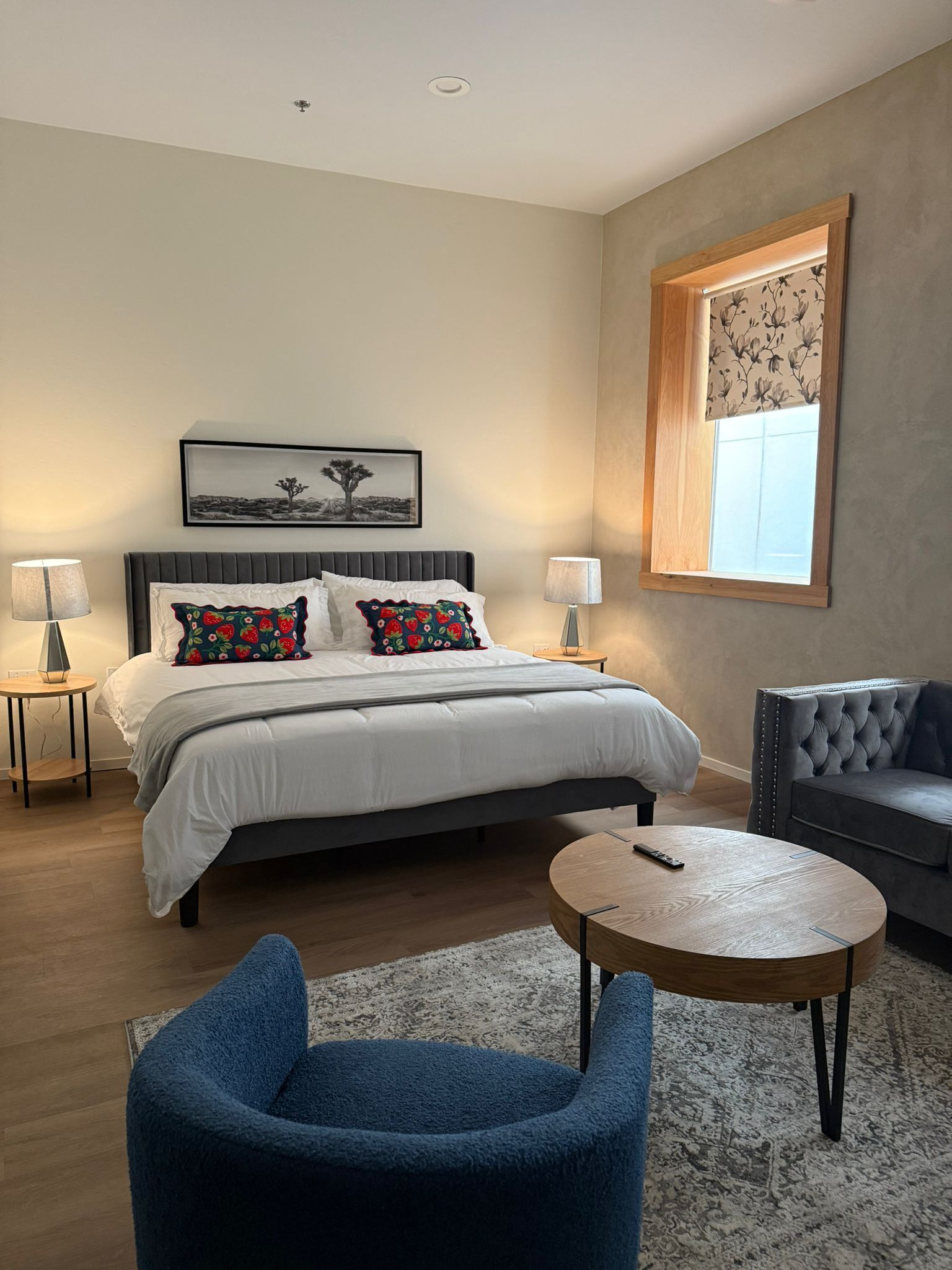 A modern bedroom with a large bed, two bedside tables, a round coffee table, a blue armchair, a tufted sofa, and a window with floral curtains.