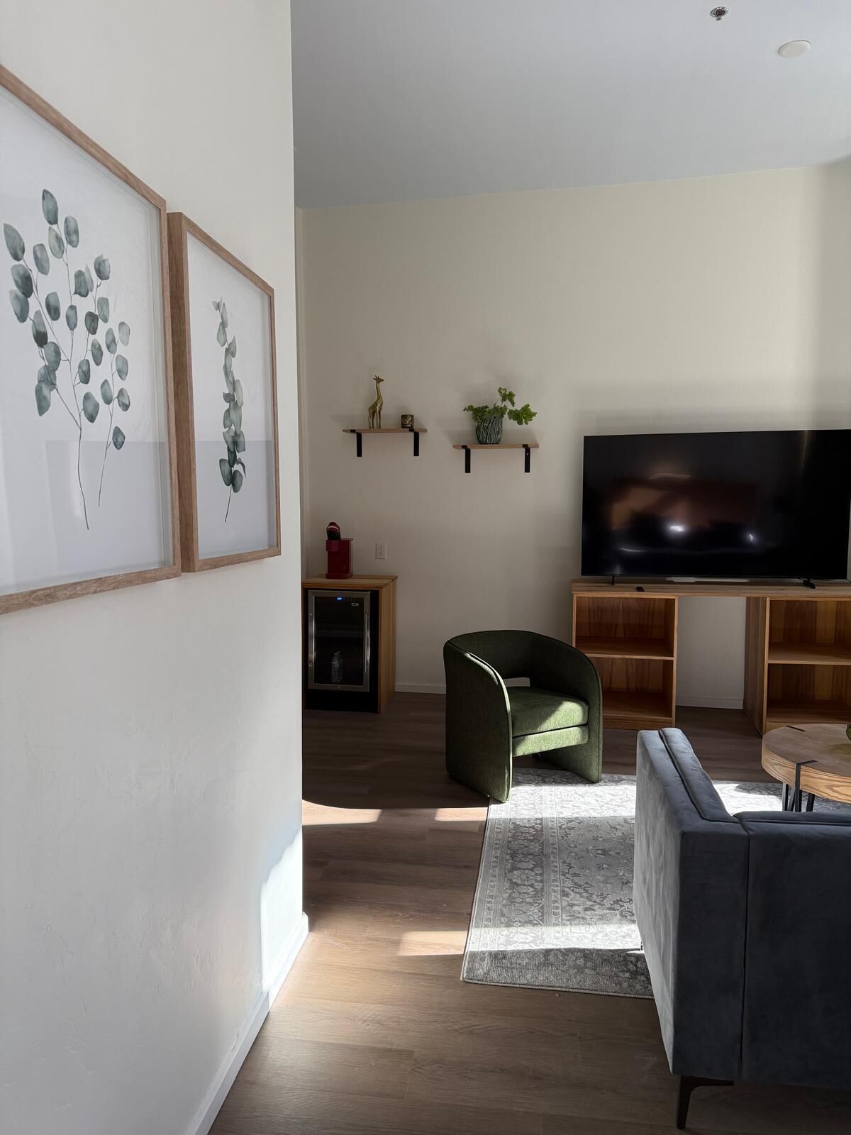 Un salon moderne avec un téléviseur à écran plat, un fauteuil vert, un canapé gris et des étagères en bois ornées d’objets décoratifs.