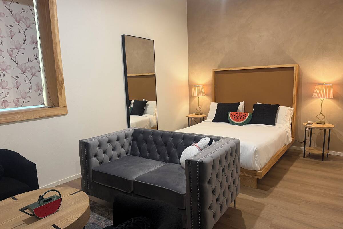 Hotellihuone, jossa on suuri sänky, napitettulla verhoilulla varustettu nojatuoli, pieni pyöreä pöytä, jonka päällä on punainen ja vihreä esine, sekä seinällä oleva peili.