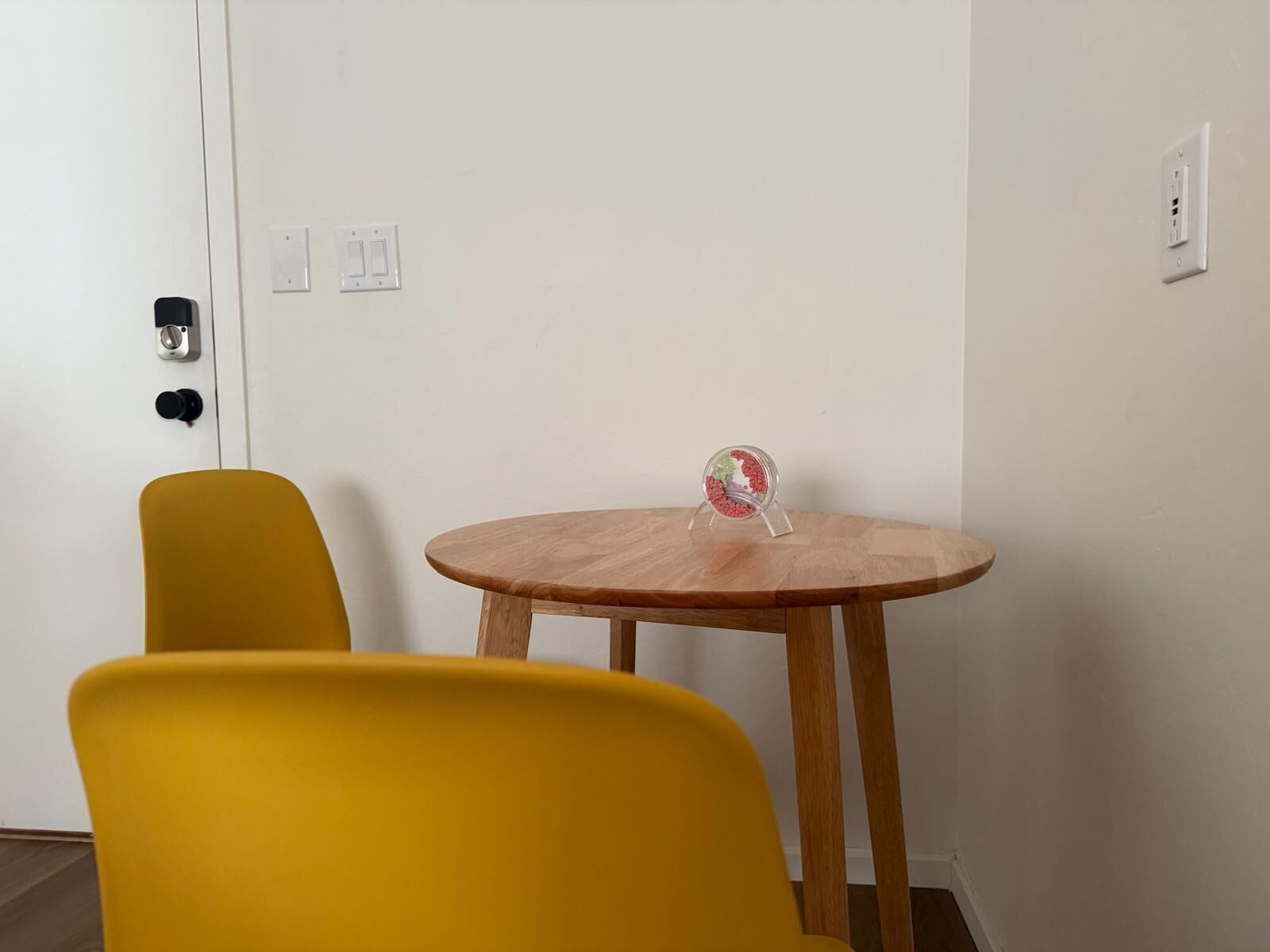 A room with a round wooden table, two yellow chairs, a small decorative item on the table, and a white door with a lock and light switch.
