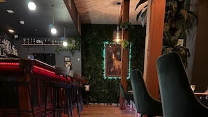 A restaurant interior with wooden flooring, green chairs, a bar area with red lighting, and a green wall with plants.
