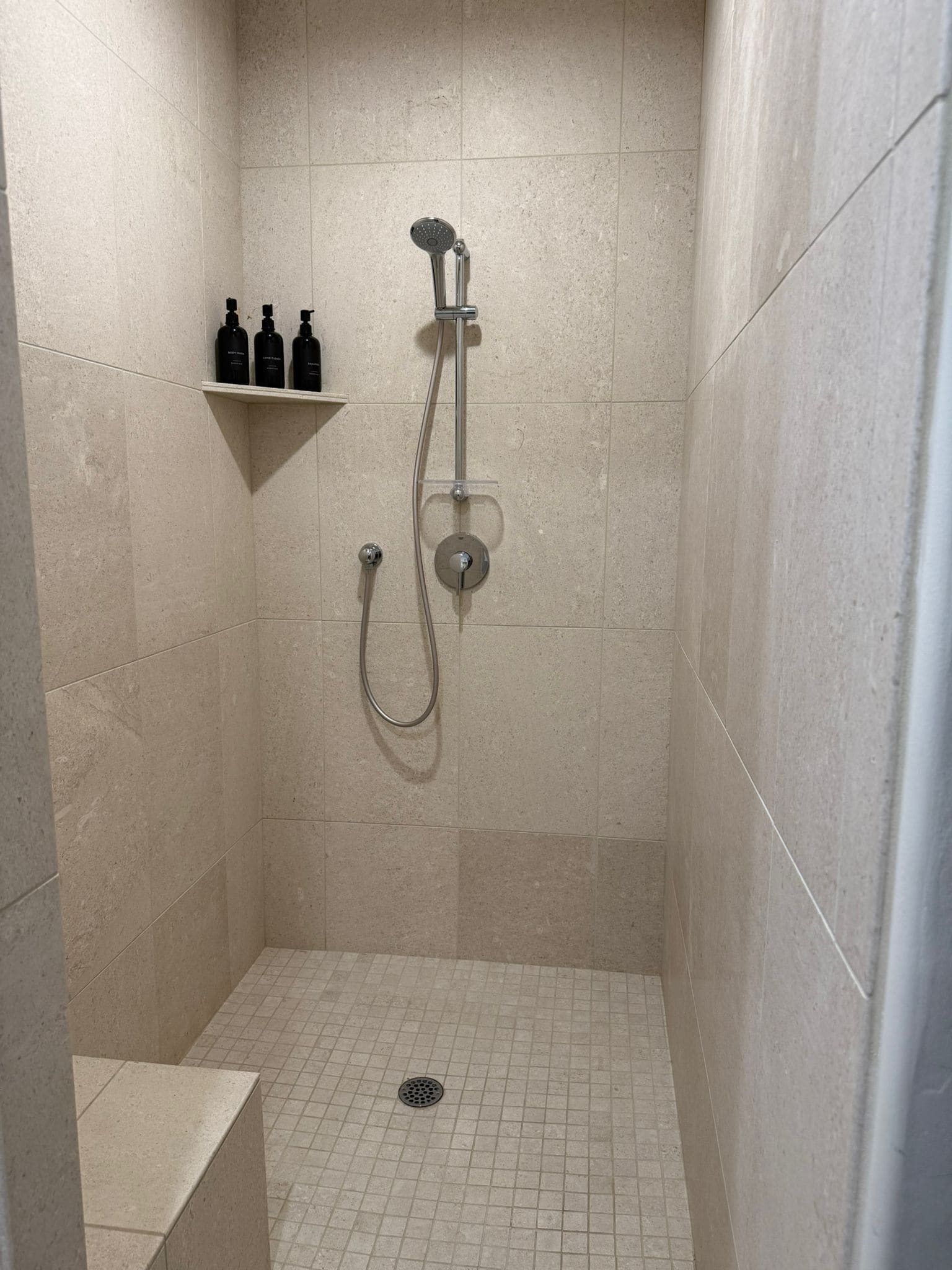 A shower with a handheld showerhead and a wall-mounted faucet, a shelf with toiletries, and a tiled floor with a drain.