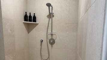 A shower with a handheld showerhead and a wall-mounted faucet, a shelf with toiletries, and a tiled floor with a drain.