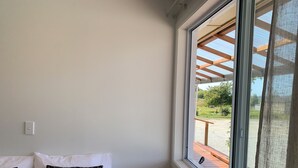 Ein Schlafzimmer mit großem Fenster, einem Bett mit weißen und gemusterten Kissen sowie Blick auf einen Garten und einen Außenbereich.