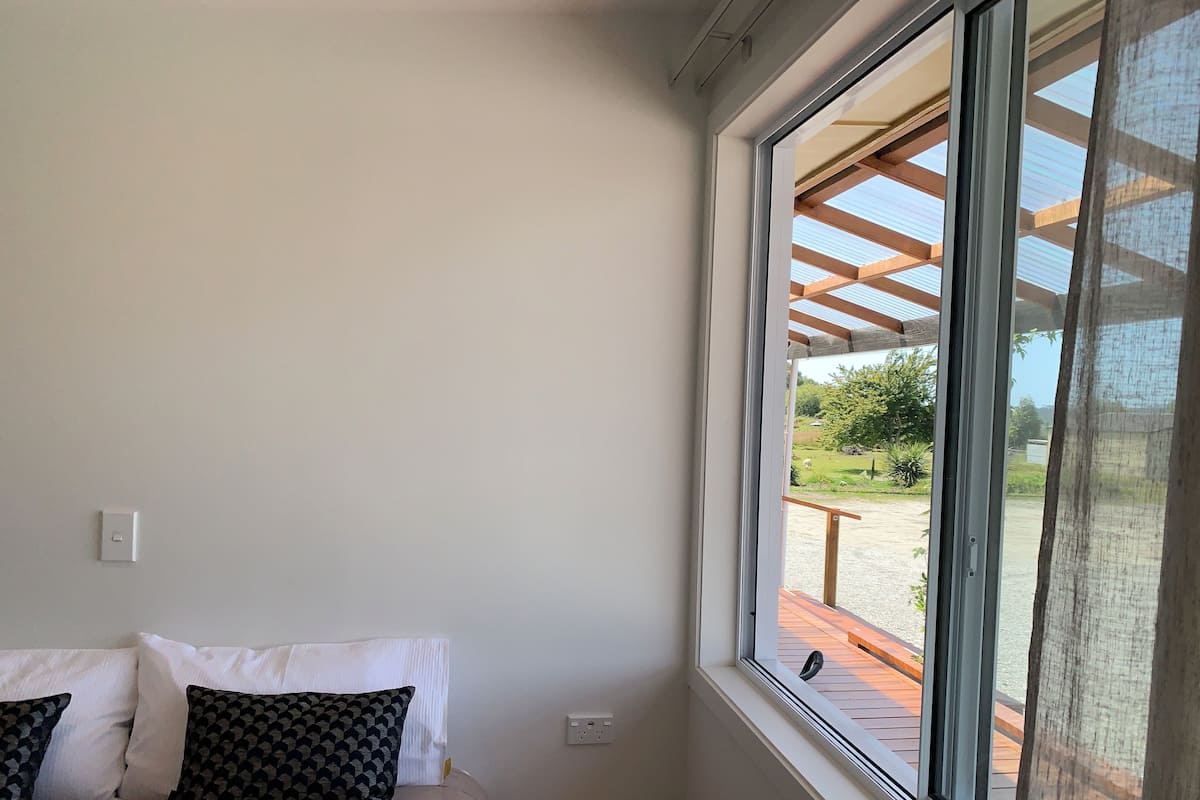 Une chambre avec une grande fenêtre, un lit agrémenté d’oreillers blancs et à motifs, et une vue sur un jardin et un espace extérieur.