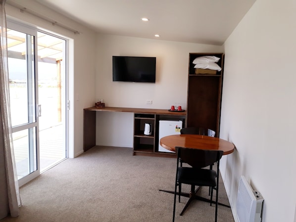 Una habitación pequeña y bien iluminada con kitchenette, un televisor de pantalla plana montado en la pared, una mesa de comedor con dos sillas y un radiador en la pared.