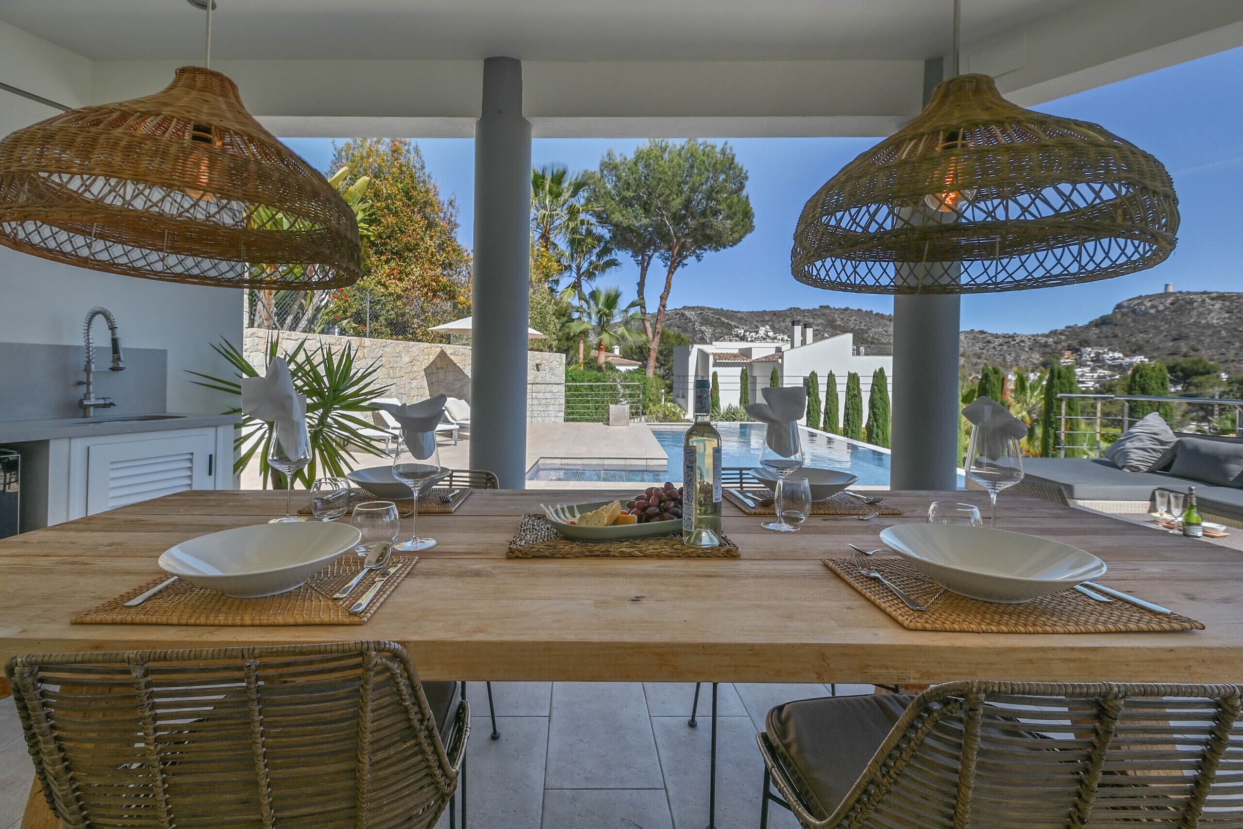 An outdoor dining area with a wooden table, wicker chairs, and a view of a pool and mountains.