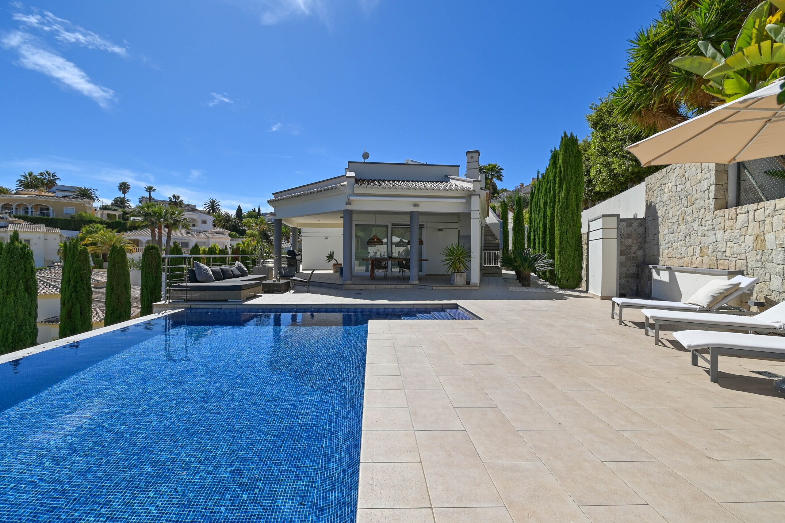 A swimming pool with lounge chairs, a stone wall, and a house in the background.