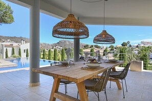 Mesa de madera preparada para comer, con lámparas colgantes de mimbre, vista a una piscina y un paisaje montañoso al fondo.