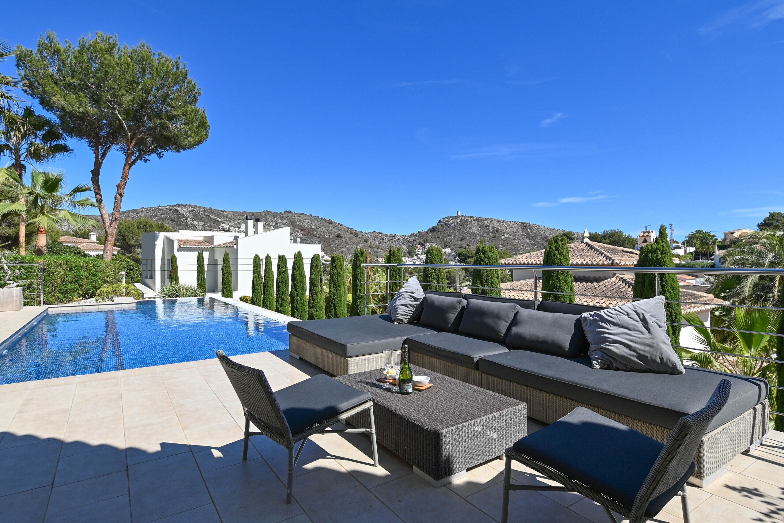 A poolside area with a sofa, chairs, and a table, overlooking a mountainous landscape.