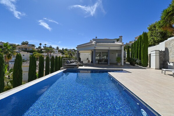Una piscina con borde de baldosas, rodeada de altos cipreses y una casa moderna al fondo.