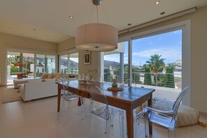 A spacious living area with a wooden dining table, clear plastic chairs, and a large window offering a view of palm trees and hills.