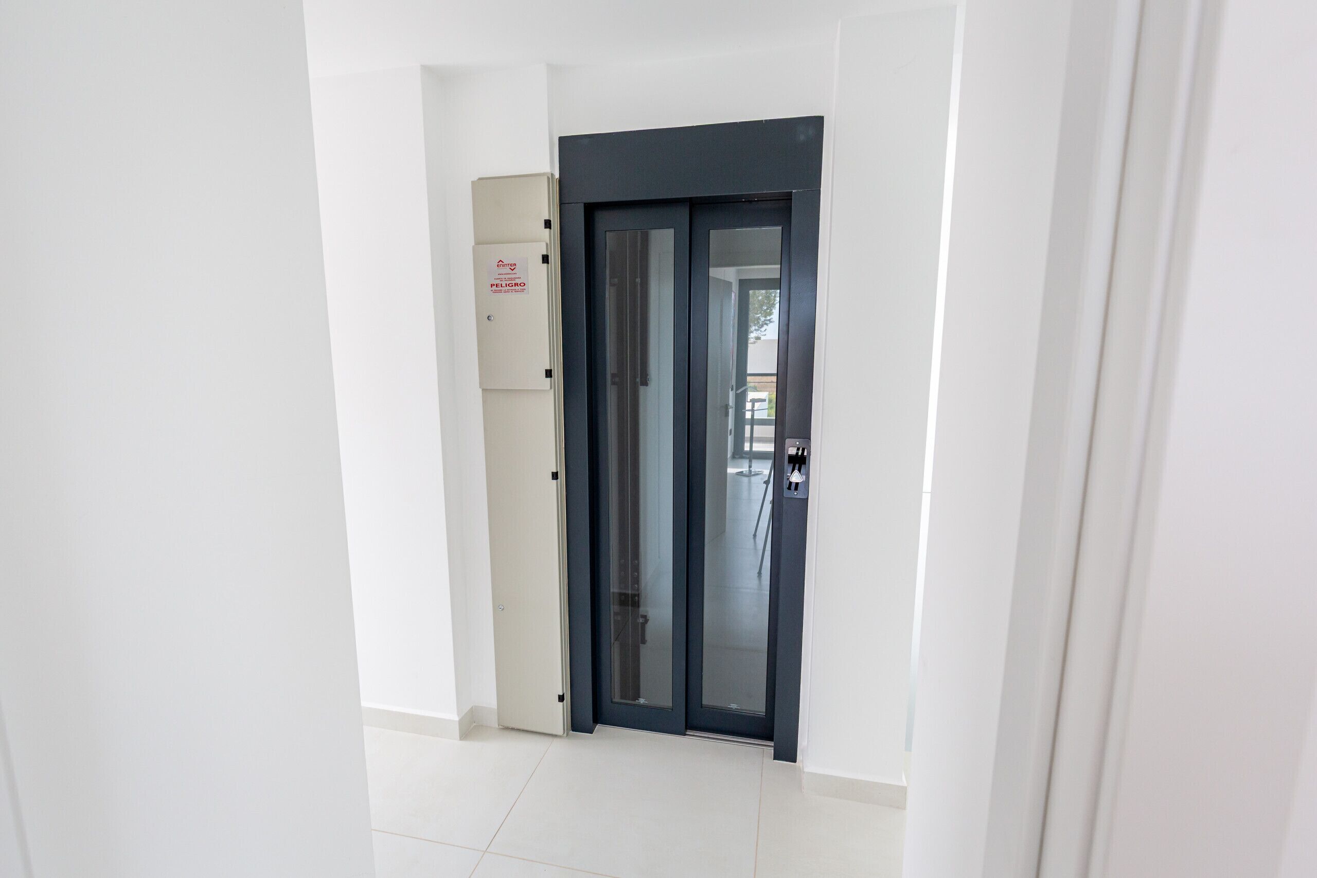 A hallway with a dark-colored door, a white wall, and a white tiled floor.