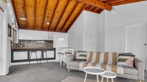 A modern kitchen with white cabinets and a wooden ceiling, a grey sofa with striped cushions, and a small round table. - Retreat on Reid Street (Merimbula)