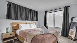 A bedroom with a wooden bed, a bedside table with a lamp, a large window with curtains, and a mirror. - Retreat on Reid Street (Merimbula)