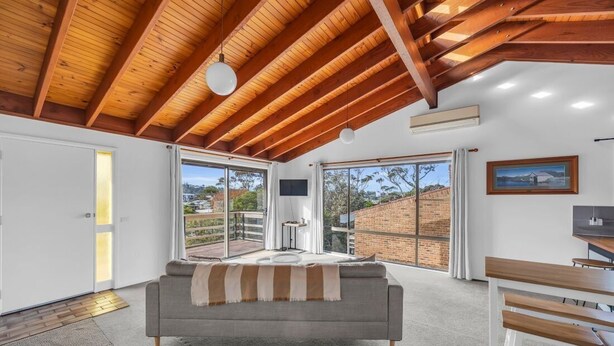 A modern interior with a high ceiling, a sofa, a dining table, and large windows offering a view of the outside. - Retreat on Reid Street (Merimbula)