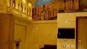 A spacious room with a high ceiling, ornate wooden columns, a large mural on the wall, a flat-screen TV mounted on the wall, and a wooden door. - Batra Guest House (Varanasi)