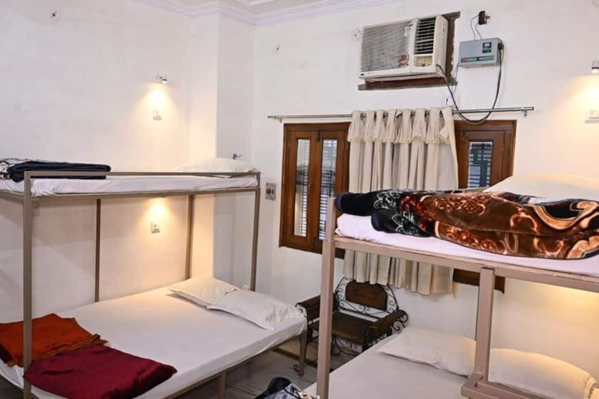 A dormitory room with bunk beds, a window with curtains, an air conditioning unit, and a small table with a chair.