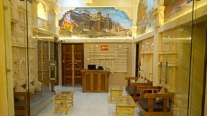 A room with traditional Indian decor, featuring carved wooden furniture, a reception desk, and a mural of a cityscape. - Batra Guest House (Varanasi)