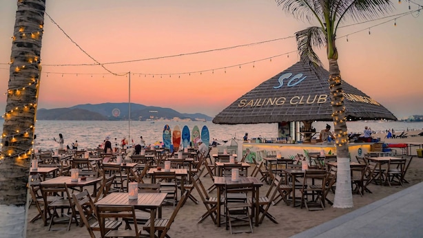 Un restaurante frente a la playa con mesas y sillas de madera, un bar con techo de paja y vistas al mar y a las montañas.