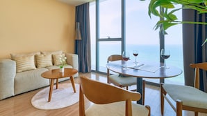 Una sala de estar moderna con un sofá, una mesa redonda y sillas, un ventanal con vistas al mar y una planta en maceta.