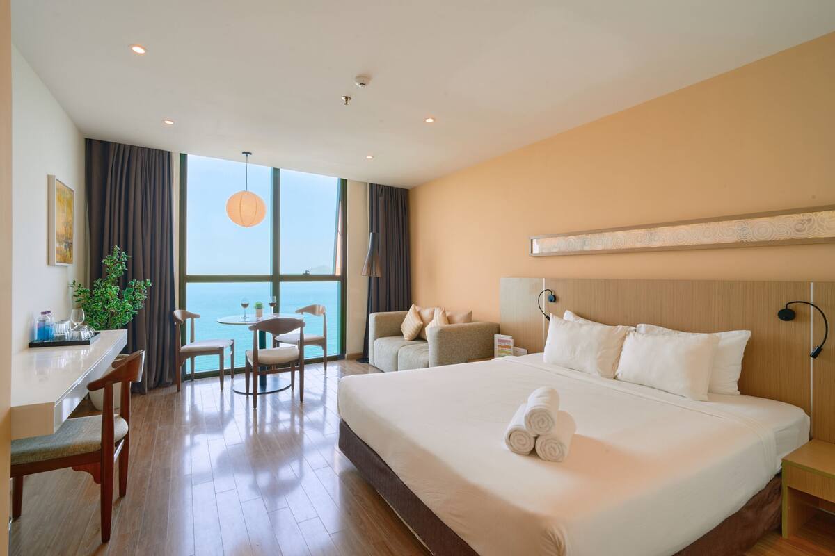 Habitación de hotel con una cama grande, un escritorio con silla, una mesita con otra silla, una planta en maceta y vistas al mar a través de un ventanal amplio.