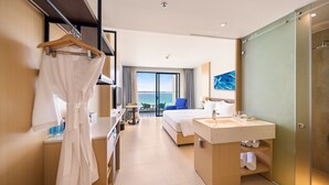 Una habitación de hotel moderna con una cama grande, un baño con lavabo y espejo, una bata colgada en un perchero y vista al mar por la ventana.