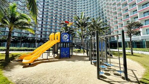 Un parque infantil con toboganes y estructuras para escalar, rodeado de palmeras y un gran edificio de hotel al fondo.