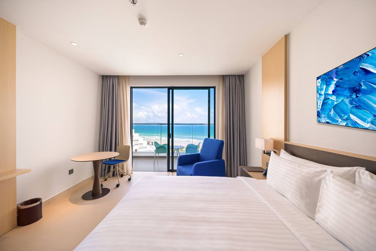 A hotel room with a large bed, a desk, a chair, and a view of the beach.