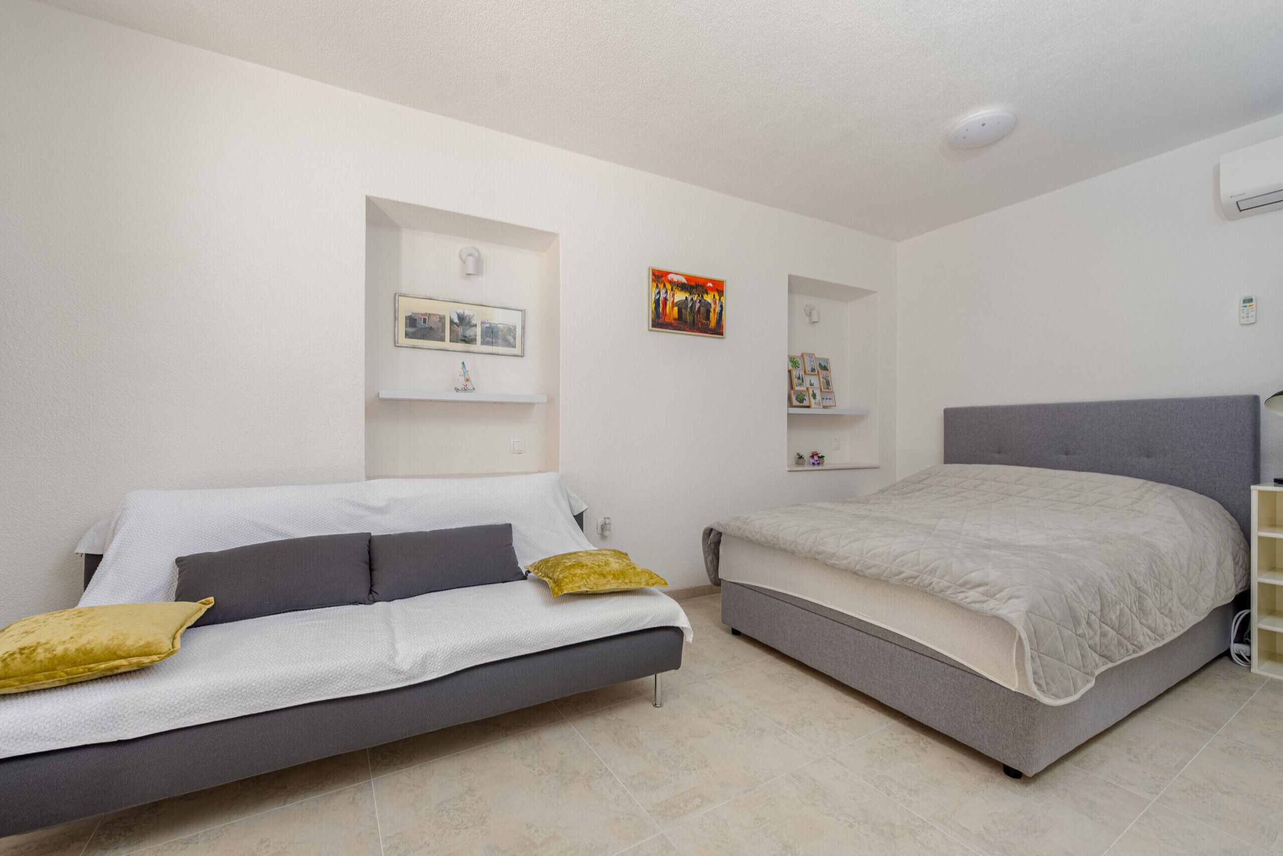 A modern bedroom with a grey sofa, a bed with a grey headboard, and a white wall with a framed picture and a small shelf.