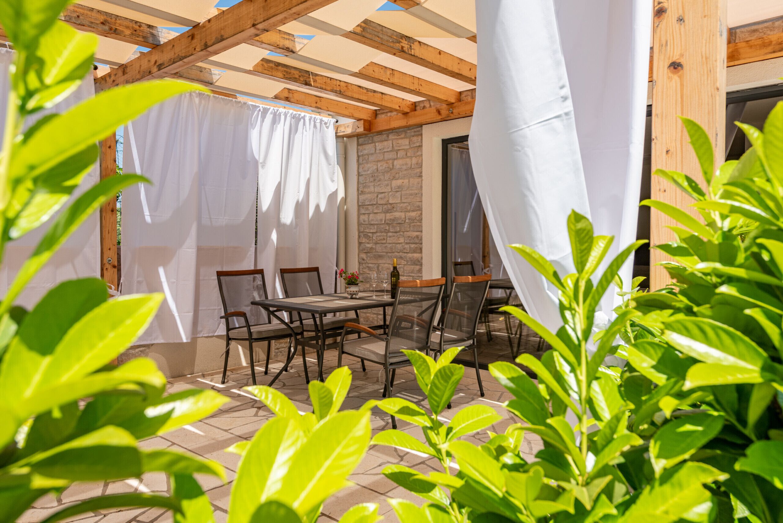 An outdoor patio with a wooden pergola, stone flooring, and a table with chairs.