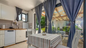 Une cuisine avec des meubles blancs, une table recouverte d’une nappe à rayures et une fenêtre avec des rideaux.