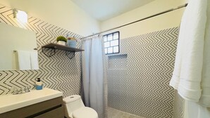 A bathroom with a white sink, toilet, and shower. There is a shelf above the sink with a plant and a bottle. The walls have a black and white zigzag pattern.