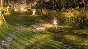 A well-lit garden with a stone pathway, lush greenery, and a seating area with a table and chairs. - Om Villas Kedungu (Tabanan)