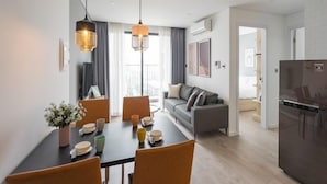 A modern kitchen with a built-in refrigerator, a dining area with a black table and orange chairs, and a living area with a gray sofa and a balcony with curtains. - Vinhomes D'Capital - SunhouseLand (Hanoi)