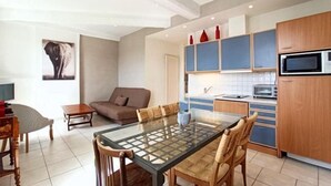 A modern kitchen with wooden cabinets, a microwave, and a dining area with a glass table and chairs. - Central 1 Bedroom Place Marche Gambetta (Cannes)
