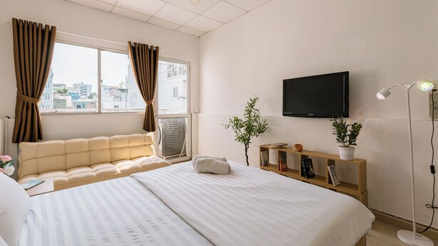 A hotel room with a large bed, a flat-screen TV mounted on the wall, a bedside table with a lamp, and a view of the city through the window. - Elysian Homestay (Ho Chi Minh City)