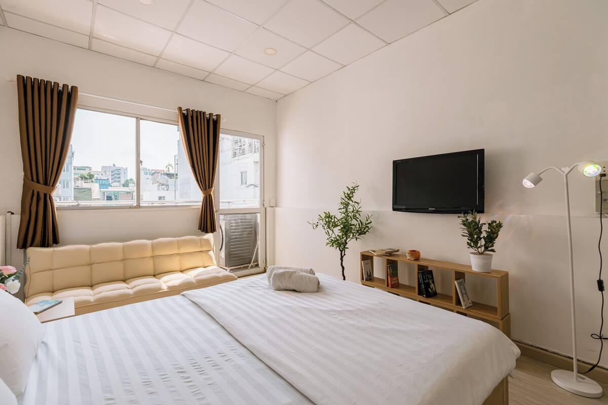 A hotel room with a large bed, a flat-screen TV mounted on the wall, a bedside table with a lamp, and a view of the city through the window.