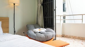 A hotel room with a bed, a bedside lamp, a checkered ottoman, and a view of a building through a window with a metal gate. - Elysian Homestay (Ho Chi Minh City)