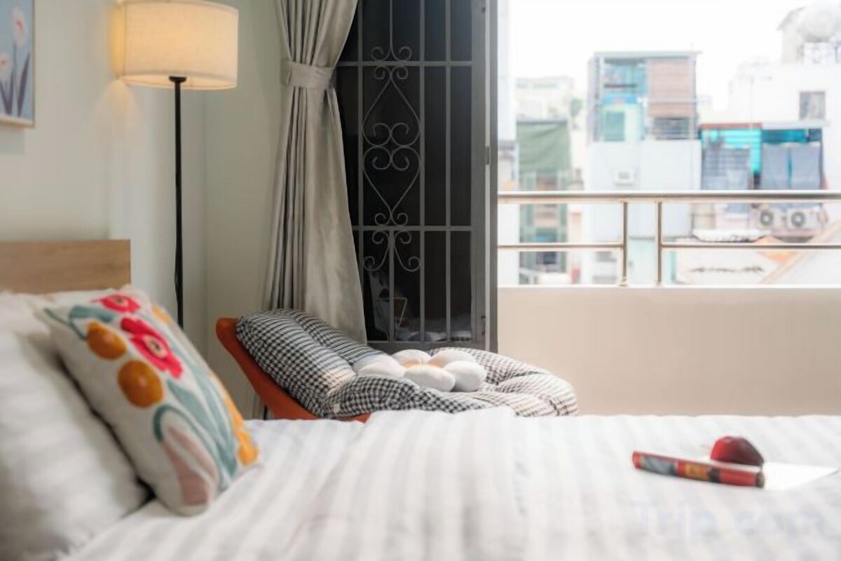 A hotel room with a bed, a chair, a window with curtains, and a view of buildings.
