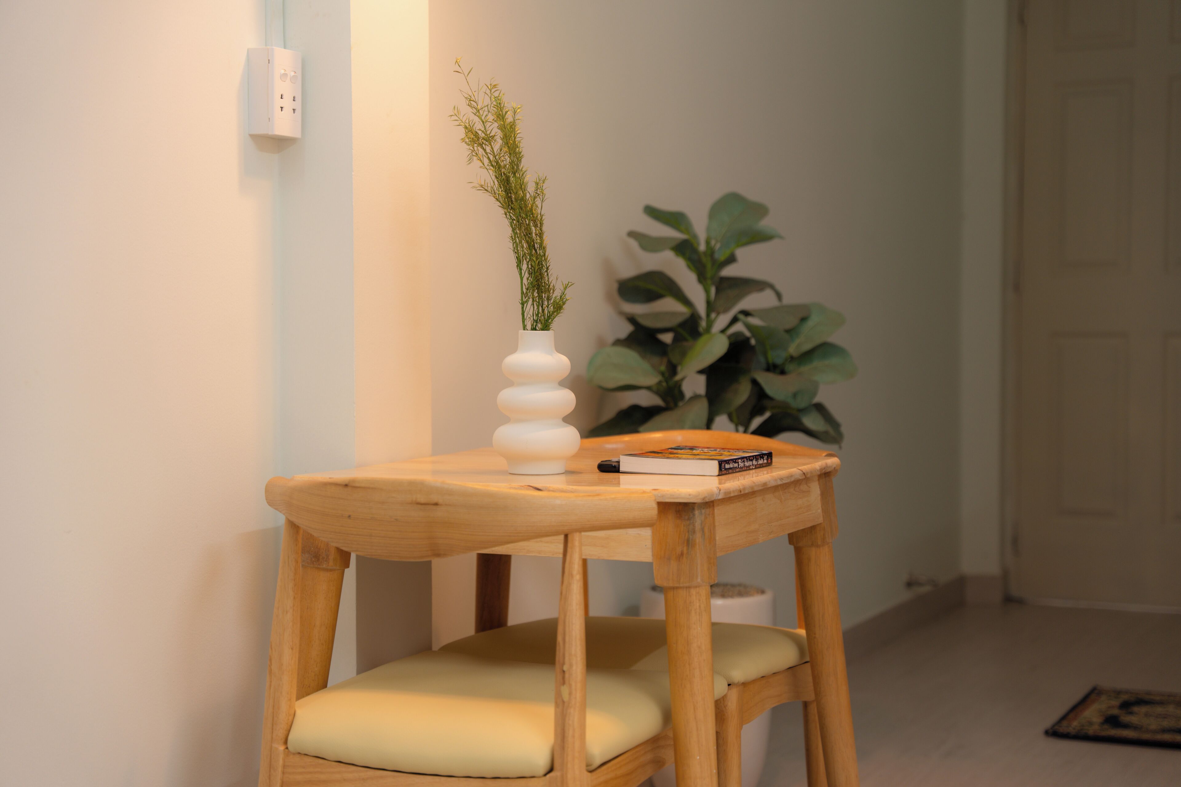 A wooden stool with a cushioned seat, a white vase with a plant, and a book on top.