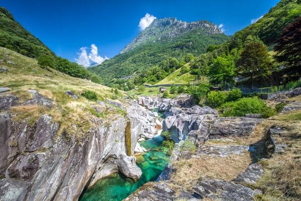 Un paisaje montañoso con un arroyo claro, terreno rocoso y un edificio pequeño al fondo.