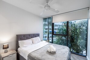 Ein modernes Schlafzimmer mit einem großen Bett, einem Nachttisch, einem Deckenventilator und Blick auf Palmen durch große Fenster.