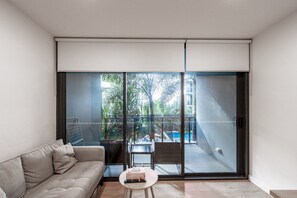 A modern living room with a sofa, a small round table, and a sliding glass door leading to a balcony with a pool and greenery.