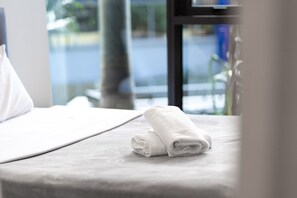 A neatly made bed with a folded white towel on it, a window with a view of a building, and a plant in the background.